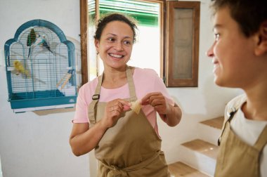 Neşeli bir anne ve çocuğu, arka planda kuş kafesi olan parlak bir mutfakta birlikte yemek pişirirken, aile bağlarını ve samimi anları vurgulayarak bir neşe anlarını paylaşıyorlar..