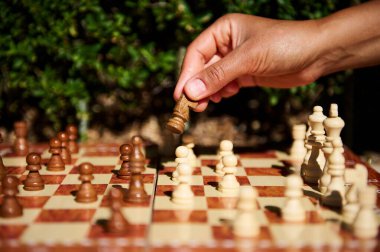 A hand holding a chess piece during a game on a wooden chessboard outdoors, promoting mental strategy and competition. clipart