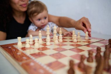 Bir anne evde satranç oynarken bebeğini sevgiyle tutar. Eğitim, boş zaman ve aile zamanı kombinasyonu, erken çocukluk gelişimi ve ebeveyn-çocuk bağlantılarını vurgulayarak büyüleyici bir şekilde resmedilir..