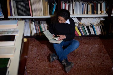 Kitaplarla dolu bir kütüphanenin zemininde okumaya dalmış öğrenme aşkı ve bilgi arayışını temsil eden bir insanın huzurlu bir anı..