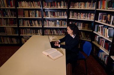 Bir kadın kütüphanede bir masada oturuyor, dizüstü bilgisayarda çalışıyor ve etrafı kitaplarla çevrili..