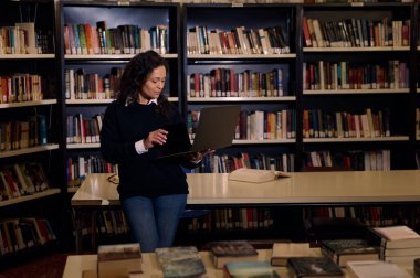 Bir kadın kütüphanede araştırma için dizüstü bilgisayar kullanıyor. Etrafında kitaplıklar ve açık ve kapalı kitaplarla dolu bir masa var. Bilgi ve odak ortamı yaratıyor..