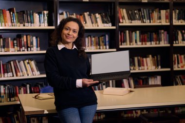 Kendine güvenen bir kadın laptopunu bir kütüphanede sunuyor, eğitim ve teknoloji entegrasyonunu iletiyor. Kitaplar ve çalışma materyalleri onu çevrelemiş, öğrenme ortamı sergiliyor..