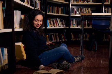 Bir kadın kütüphane zemininde oturuyor, etrafı kitaplarla çevrili, okuyor ve dizüstü bilgisayar kullanıyor. Atmosfer, geleneksel okuma ve modern teknolojiyi birleştirerek konsantrasyon, öğrenme ve entelektüel uğraşlar taşır..