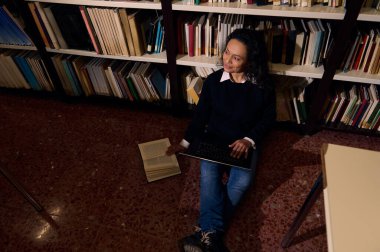 Kitaplarla çevrili bir kütüphanenin zemininde oturan bir kadın çalışmak için dizüstü bilgisayar kullanıyor. Sahne, eğitim, okuma, araştırma ve sessiz ve huzurlu bir ortamda bilgi arayışını temsil ediyor..