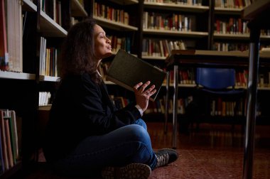 Eserlerle dolu raflarla çevrili bir kitap okumaya dalmış bir kadın kütüphanede rahatça oturuyor..