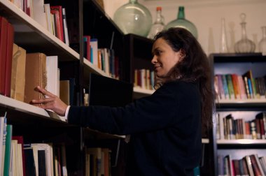 A woman is seen browsing books on a library shelf, exuding curiosity and engagement with the environment. The books and warm atmosphere emphasize a love for knowledge and intellectual exploration. clipart