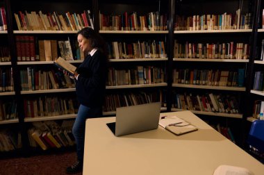 İyi düzenlenmiş bir kütüphanede kitap raflarıyla kitap karıştıran bir kadın bilgiye ve öğrenmeye dikkat çekiyor. Yakınına bir dizüstü bilgisayar ve defter yerleştiriliyor. Araştırma veya çalışma öneriliyor..