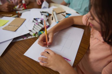 Genç bir çocuk sanat eserleri yaratmaya dalmışken kalem ve kağıt kullanıyor. Çeşitli sanat malzemeleri, yaratıcı ve eğlenceli bir eğitim ortamında öğrenmeyi gösteren bir tablo üzerine dağılıyor..