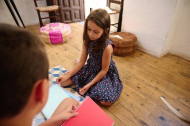 Okul projesinde işbirliği yapan iki çocuk, ahşap zeminde, köy ortamında oturuyorlar..