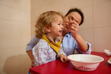 Joyful moment of a mother feeding her happy child in a warm kitchen setting. The child eagerly enjoys the meal, capturing a warm bonding moment highlighting joy and love. clipart