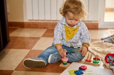 Eğitici oyuncakların yanında yerde oturan küçük bir çocuk, parlak bir oturma odasında bilişsel ve motor becerilerini gösteriyor..