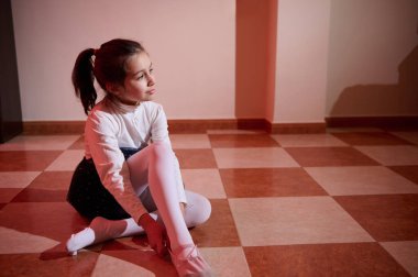 Bale kıyafeti giyen bir kız sıcak ışıkla aydınlatılmış fayanslı bir zeminde oturur, sakinlik ve yansıma anını yakalar..