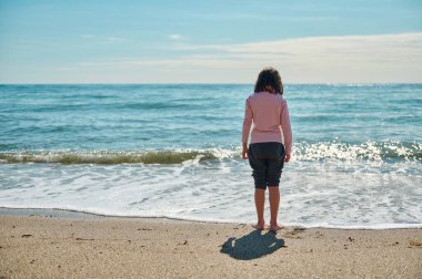 Sahilde çıplak ayakla duran genç bir kız, ılık yaz güneşinin altında sakin okyanusa bakıyor, huzur ve sükuneti çağrıştırıyor..