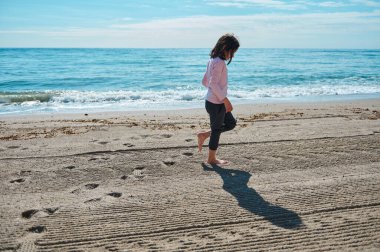 Genç bir çocuk, okyanusun kenarındaki pürüzsüz kumlu bir sahilde çıplak ayakla yürüyor. Parlak, güneşli bir gökyüzü altında. Doğal neşe ve yazın özünü somutlaştırıyor..