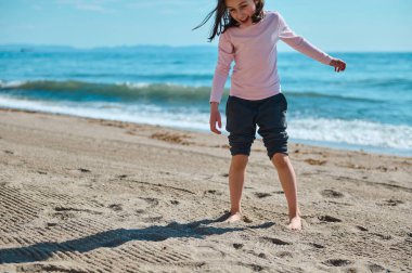 Neşeli bir kız okyanus kıyısında kumlu sahilde yalınayak dolaşır, güneşli günün tadını gülerek ve oynayarak çıkarır. Deniz dalgaları manzarayı güzelleştirir..