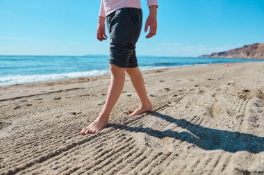 Açık gökyüzü ve manzaralı kumlu sahilde yalınayak yürüyen bir insan, rahatlama ve doğayla bağlantı hissi uyandırıyor..