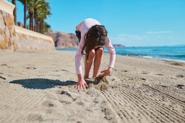 Genç bir kız kumla oynarken, okyanusun yakınındaki kumlu bir sahilde sıcak ve güneşli günün tadını çıkarırken görülüyor..