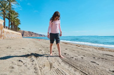 Genç bir kız kumsalda güneşli bir günün tadını çıkarıyor. Huzurlu okyanusun yanındaki yumuşak kumda yalınayak yürüyor..