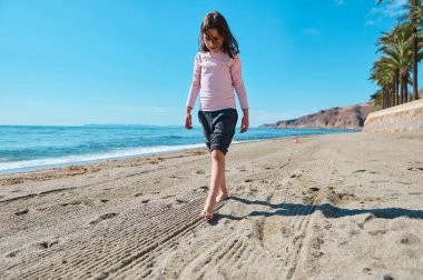 Kumlu bir sahilde çıplak ayakla yürüyen çocuk manzaralı okyanus ve palmiye ağaçları altında canlı güneşli bir gökyüzünün altında.