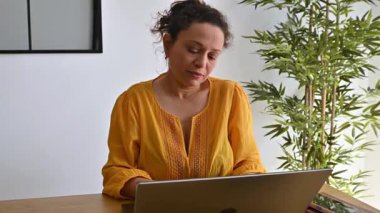 Sarı gömlekli kadın evde çalışıyor. Düşünmek için bir dakikasını ayırıyor ve laptoptan uzağa bakıyor..