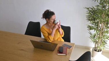 Sarı elbiseli kadın laptopuyla çalışmaya başlamadan önce kahvesini yudumluyor..