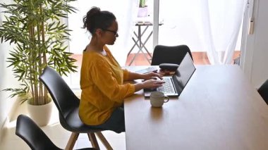 Evde dizüstü bilgisayarıyla çalışan bir kadın. Yemek masasında oturuyor..