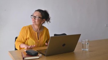 Bir kadın dizüstü bilgisayarında çalışıyor, ara sıra etrafa bakıyor ve konuşuyor. Ara verir, kollarını kavuşturur ve çalışmaya devam eder..