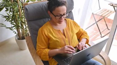 Evden çalışan bir kadın. O rahat bir sandalyede oturuyor ve dizüstü bilgisayarında yazıyor..