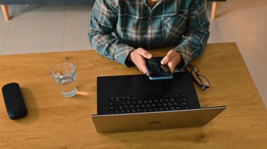 Bilgisayarlı, akıllı telefonlu, gözlüklü ve su bardağıyla ahşap bir masada çalışan birinin tepeden tırnağa görüntüsü, günlük ev ortamında çoklu görev ve modern dijital iletişimi tasvir ediyor..