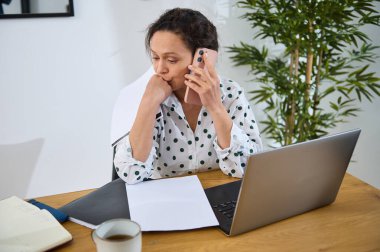 Evden uzakta çalışan, dizüstü bilgisayar ve telefonla birden fazla iş yapan genç bir kadın. Günlük çalışma alanı bir fincan kahve içeriyor, rahat ve verimli bir uzaktan çalışma ortamı sunuyor..