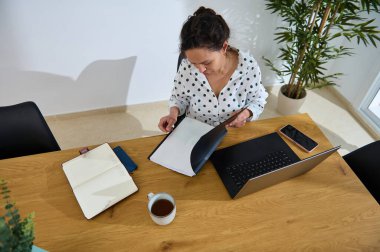 Genç bir kadın, ev ofisinde, modern uzaktan çalışma ve organizasyonu gösteren bir dizüstü bilgisayar, kahve ve defterlerle ahşap masasındaki belgeleri inceliyor..