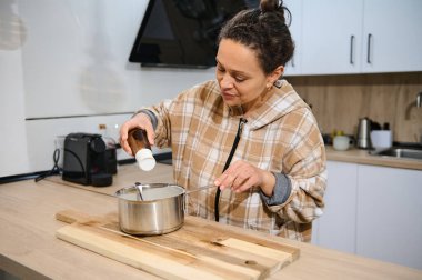 Bir kadın şık bir mutfakta yemek pişirirken tencereye malzeme koyarken görülüyor..