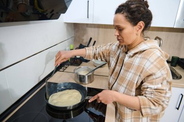 Çağdaş bir mutfak sobasında yapışmaz tavada krep pişiren bir kadın. Hoş ve düzenli bir ev ortamında rahat bir atmosfer yayarak hazırlanmaya odaklanmış durumda..