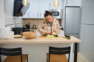 Genç bir kadın sıcak, modern bir mutfakta taze malzemelere ve ev yapımı yemeklere odaklanmış sağlıklı bir salata hazırlıyor. Yaşam tarzı, sağlık ve mutfak faaliyetlerini desteklemek için ideal.
