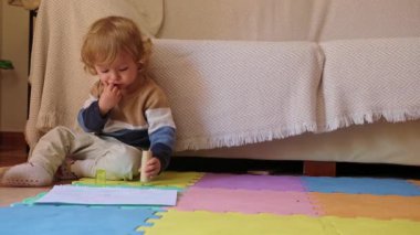 Şirin çocuk renkli bir oyun hasırının üzerinde oturur ve fosforlu kalemi keşfeder. Meraklı çocuk oturma odasındaki kanepenin yanında oturur. Bebek kalemi ağzına koyuyor..