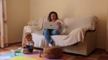 Bir anne, bebeğine göz kulak olurken uzaktan bilgisayarının başında çalışır ve rahat bir evde yerde oyuncaklarla oynar..