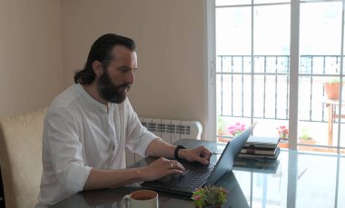 Sıcak ev dekoru ve doğal ışıklandırmayla çevrili cam bir masada dizüstü bilgisayarıyla çalışan sakallı bir adam. Modern uzak çalışma ve samimi ortamların özünü yakalar.