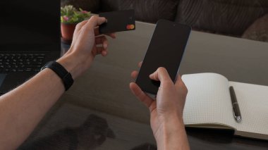 Bir kişi, çalışma alanı gereksinimleriyle çevrili, modern teknoloji ve iş kolaylığını vurgulayan bir kredi kartıyla akıllı telefon kullanmaktadır..