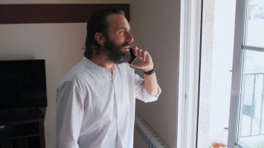 Bir pencerenin yanında duran genç adam, odaklanmış bir ifadeyle akıllı telefonuyla konuşuyor. Beyaz bir gömlek giyiyordu. Ofis ortamında iş gibi görünüyordu..
