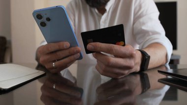 Çevrimiçi bankacılık ve alışveriş yaparken elinde akıllı telefon ve kredi kartı olan bir adamın yakın plan görüntüsü. Modern finans teknolojisi ve e-ticaret kavramlarını tasvir etmek için ideal..