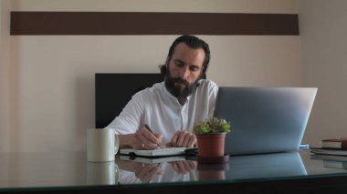 Dizüstü bilgisayarı, kupası ve saksı bitkisi olan bir masada sakallı profesyonel yazma notları..