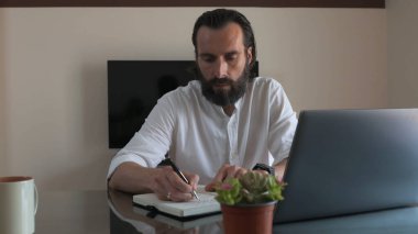 Masalarında çalışan bir profesyonel, dizüstü bilgisayar kullanırken deftere yazı yazıyor. Modern ortamlarda üretkenlik, uzak çalışma, yaratıcı düşünme veya serbest çalışma temaları için uygundur..