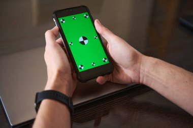 Akıllı bir kaset takan, elinde yeşil ekran gösteren bir akıllı telefon tutan biri. Akıllı telefon ahşap bir masanın üzerindeki dizüstü bilgisayarın üzerinde duruyor. Teknoloji ve tasarım uygulamalarını öneriyor..