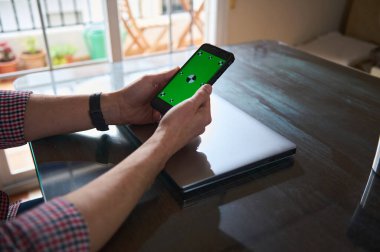 Adam, kapalı bir masada oturan yeşil ekranı gösteren bir akıllı telefonla etkileşime giriyor. Yakınlarda, çalışma alanını gösteren parlak bir dizüstü bilgisayar masanın üzerinde duruyor. Doğal ışık odayı dolduruyor..