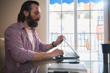 Sakallı bir adam bir masada dizüstü bilgisayar kullanarak oturur. Sahne, doğal ışık ve üretkenliğe olanak sağlayan rahat bir ortamda düşünceli bir çalışma ortamı tasvir ediyor..