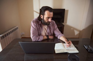 Genç bir profesyonel dizüstü bilgisayarla çalışırken bir deftere yazı yazarak bir proje planlamaya odaklanır..