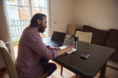 Bir adam rahat bir ev ortamında cam bir masada oturuyor, yanında bir dizüstü bilgisayarla bir şeyler yazıyor, verimliliği vurguluyor ve uzaktan çalışma düzenine odaklanıyor..