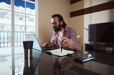 Bir iş projesine odaklanan, parlak bir ev ofisinde not alırken dizüstü bilgisayar kullanan bir serbest çalışan..