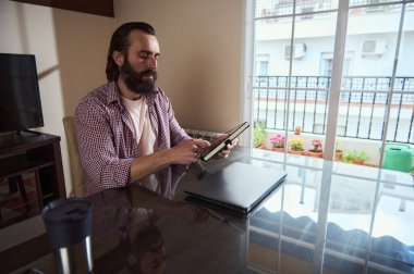 İyi aydınlatılmış bir odada oturan bir adam çalışma alanındaki dijital tabletle yanında bir dizüstü bilgisayarla oturuyor. Ayarlar, uzak çalışma için ideal konforlu ve üretken bir ortam olduğunu gösteriyor..